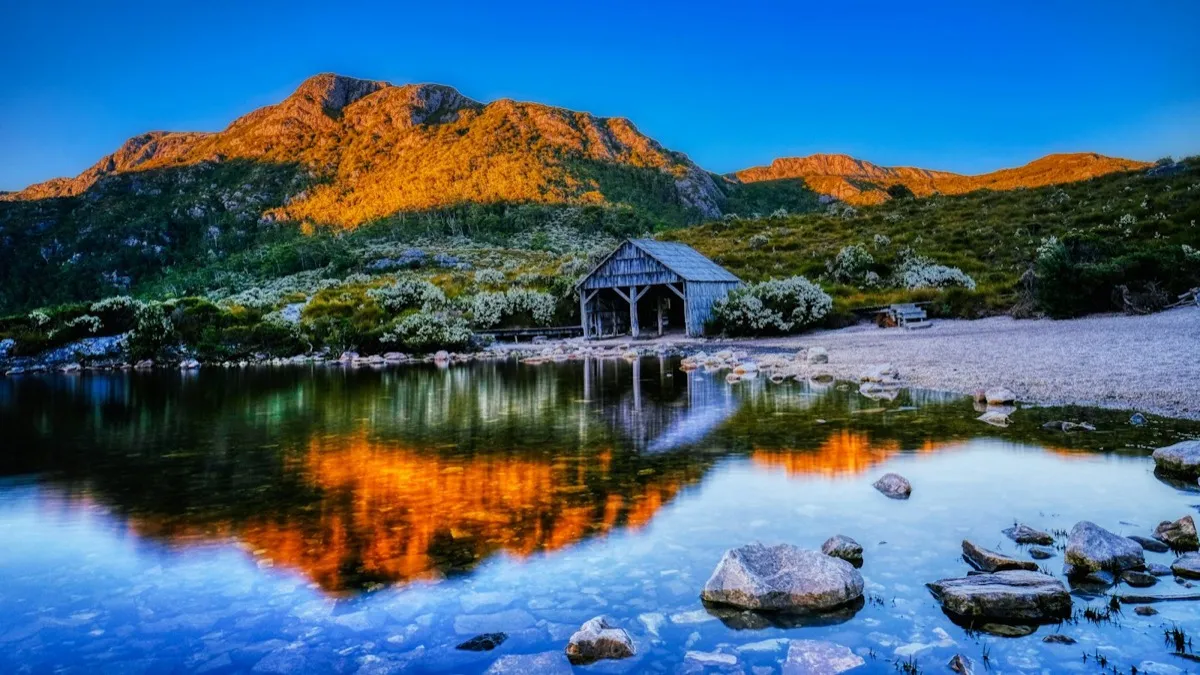 A timber boatshed on a still lake below a mountain lit orange at sunrise